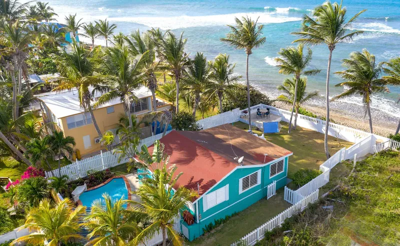 Aerial view of a colorful beachfront home with palms, a white fence, and a pool